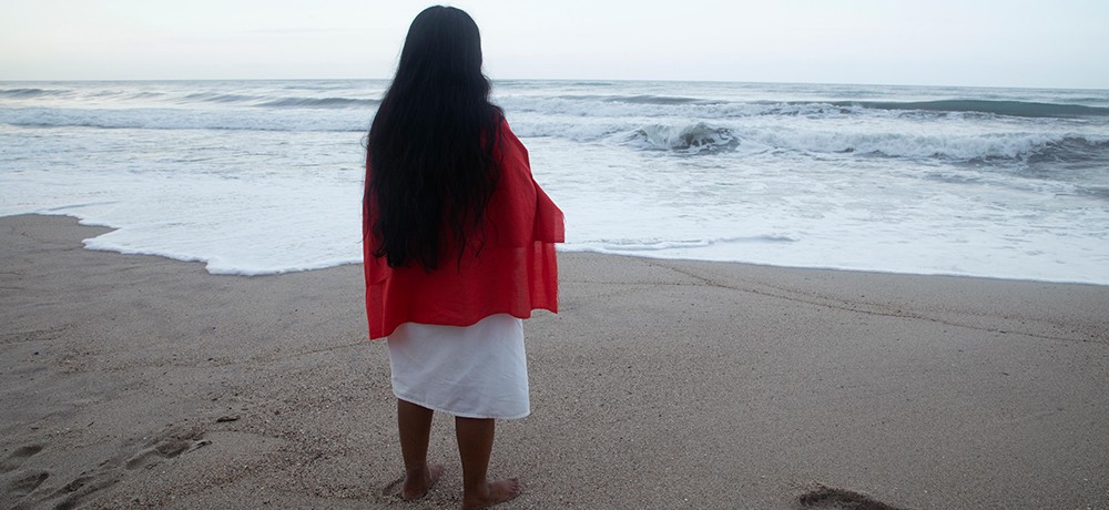 A woman stands on a sandy beach with her back to the camera; she is wearing a white dress with a bright red shawl draped over her shoulders.