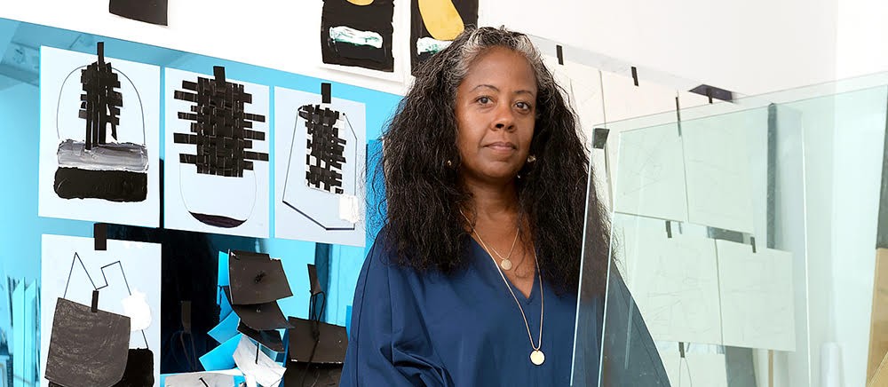 The artist, Torkwase Dyson, a woman in a blue shirt with long black hair, standing in her art studio in front of her artwork.