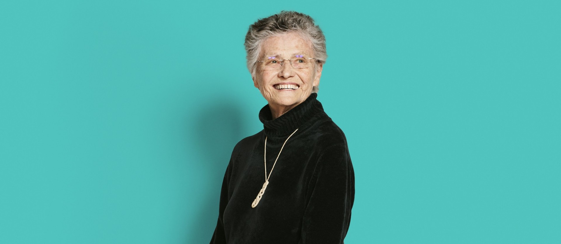 Musician Annea Lockwood: Woman with gray hair and glasses with black shirt with silver necklace in front of a turquoise-colored background.
