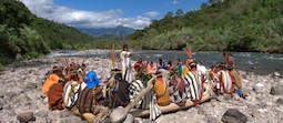 About a dozen people dressed in colorful tribal costumes sitting on a rocks and logs near a river with mountains in the background.