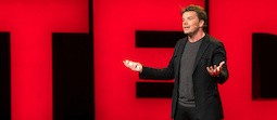 Architect Bjarke Ingels, brown hair, dressed in dark colors, speaking on a stage with red neon letters, spelling TED, behind him.