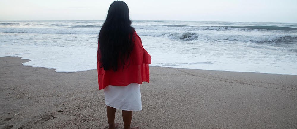 Woman with long black hair, wearing red shawl and white dress, with her back to camera looking out to the sea.
