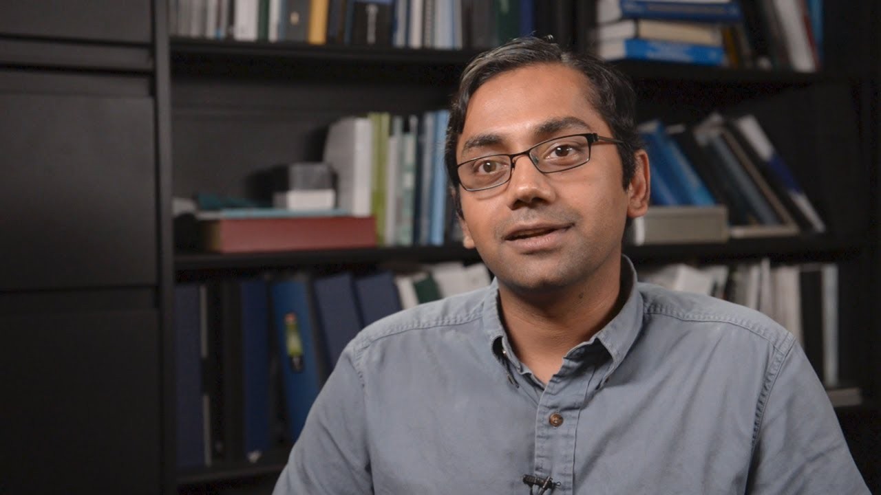 Kartik Chandran, man with glasses talking into the camera, with a shelf of books behind him.