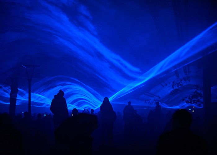 Roughly a dozen people walk through an outdoor space illuminated with blue laser light.