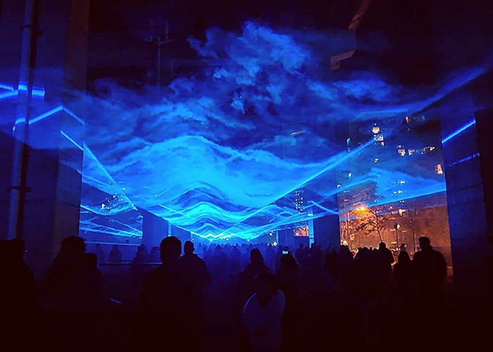 Dozens of people wander through an outdoor space illuminated by blue lasers that create the effect of walking through water.