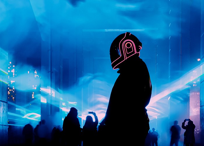 A person wearing a helmet lined with laser LED lights stands in an outdoor space illuminated by blue laser light.