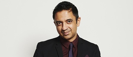 A man in a dark suit and tie sits on a stool, looking downward slightly and smiling into the camera.