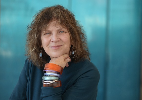 A woman, wearing a dark teal dress and standing in front of a sea blue background, rests her head on her hand and smiles into the camera.