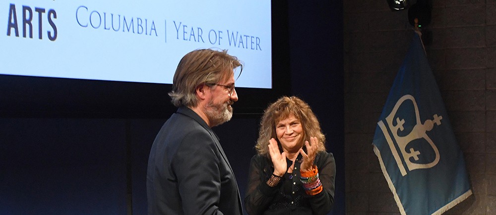 Bearded man being applauded on a stage by a woman with curly hair.