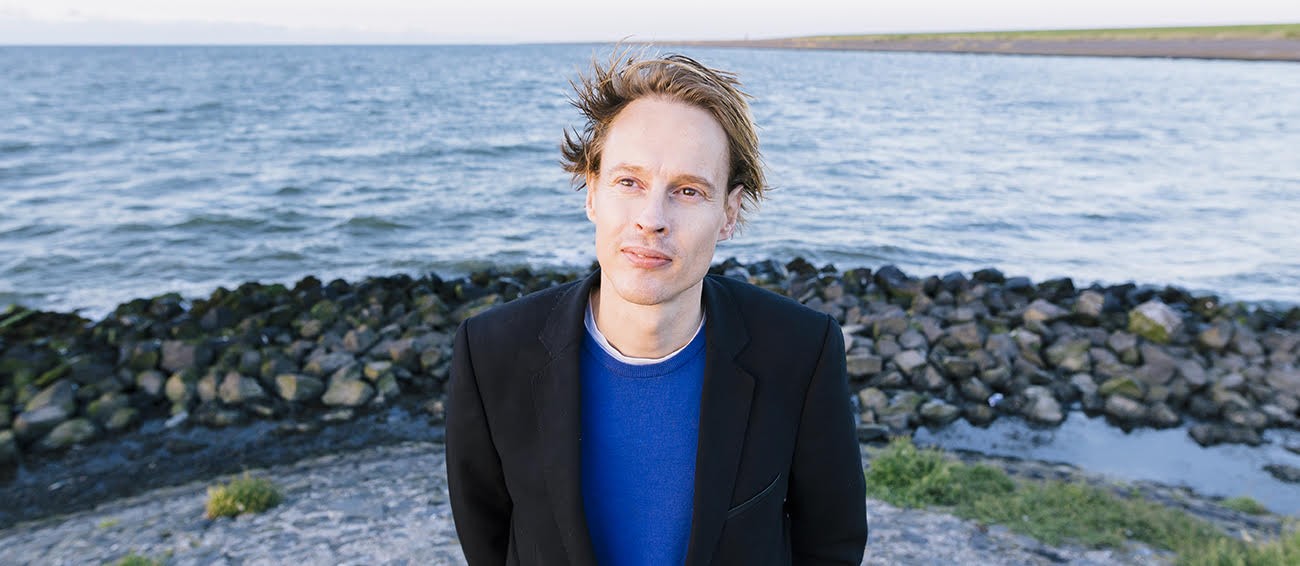 A man with blue shirt and navy blue blazer standing in front of a body of water.