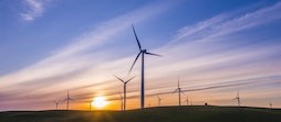 A dozen solar-powered windmills stretched across a barren field with sunsetting in the distance.