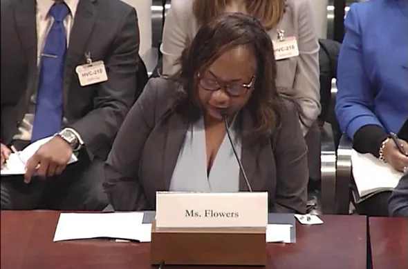 A woman in a dark gray suit sits at a table speaking into a microphone, with rows of people seated behind her taking notes.