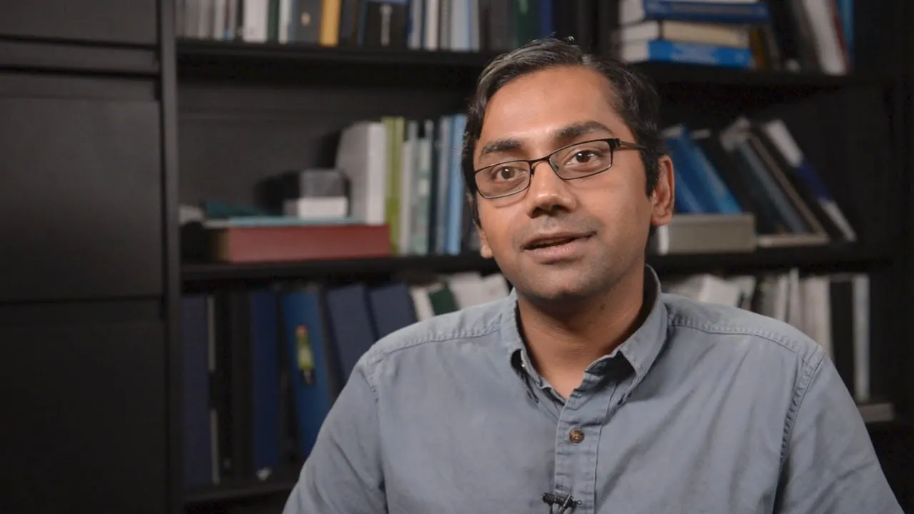 Kartik Chandran, man with glasses talking into the camera, with a shelf of books behind him.