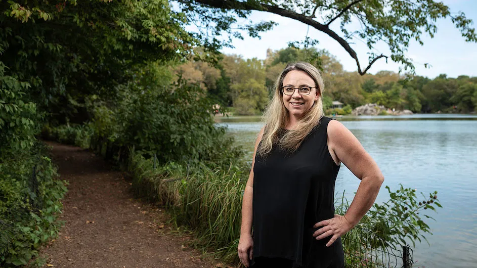 Woman with long blonde hair and glasses standing in front of a body of water.
