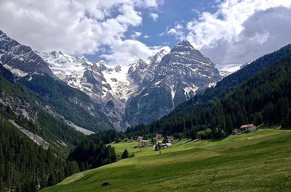 A photograph of glaciers on mountaintops in the far distance beneath blue skies scattered with clouds, a verdant valley covered with green trees and pasture